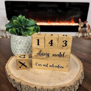 May include: Wooden countdown blocks with the numbers 1, 4, 3, and 0, along with the text "days until" and "Our next adventure". The blocks are arranged in a stack on a wooden slice.