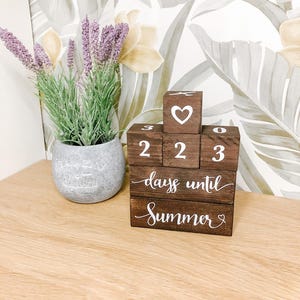 May include: A wooden block calendar displaying the numbers 2, 2, and 3. The top block features a heart. The bottom block reads "days until Summer" in white script. A potted lavender plant is to the left.