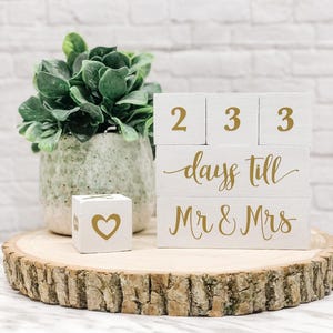 May include: White wooden blocks with gold lettering spelling out "233 days till Mr & Mrs" on a wooden slice. A small white block with a gold heart is in the foreground.