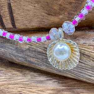 Puede incluir: Un collar de cuentas rosa y blanco con un dije de concha dorada y una perla en el centro.