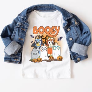 A white t-shirt with a Halloween-themed graphic design featuring cartoon characters such as Booyie, a ghost, and a skeleton, along with jack-o-lanterns and a cemetery scene.
