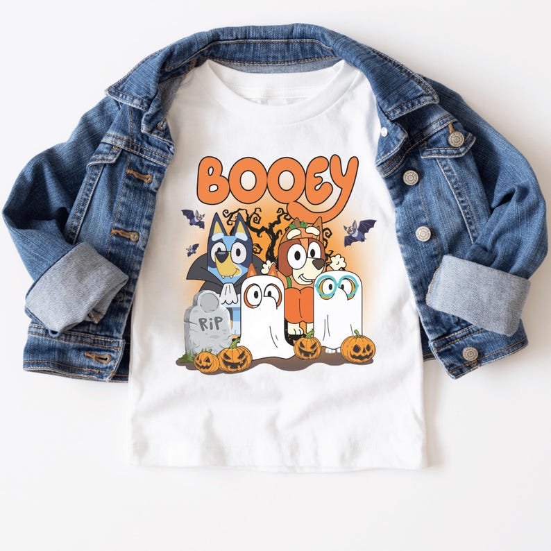 A white t-shirt with a Halloween-themed graphic design featuring cartoon characters such as Booyie, a ghost, and a skeleton, along with jack-o-lanterns and a cemetery scene.
