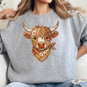 May include: A heather gray crewneck sweatshirt featuring a central graphic of a Highland cow. The cow is depicted with a patchwork-style design in shades of orange, brown, and cream, wearing a patterned bandana around its neck with autumn leaves.