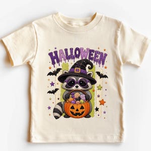 May include: A cream-colored short-sleeve t-shirt featuring a graphic of a raccoon wearing a purple witch hat and glasses, holding a jack-o'-lantern bucket filled with candy. The word HALLOWEEN is printed in purple above the raccoon.