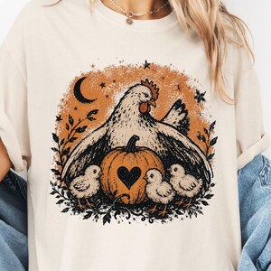 May include: A cream-colored short-sleeve t-shirt featuring a graphic print of a hen with three chicks and a pumpkin. The pumpkin has a heart-shaped cutout. The design includes a crescent moon, stars, and foliage in shades of orange and black.