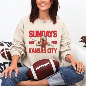 A woman sitting on a couch wearing a sweatshirt with a graphic of a Native American warrior and the text "SUNDAYS ARE FOR KANSAS CITY".