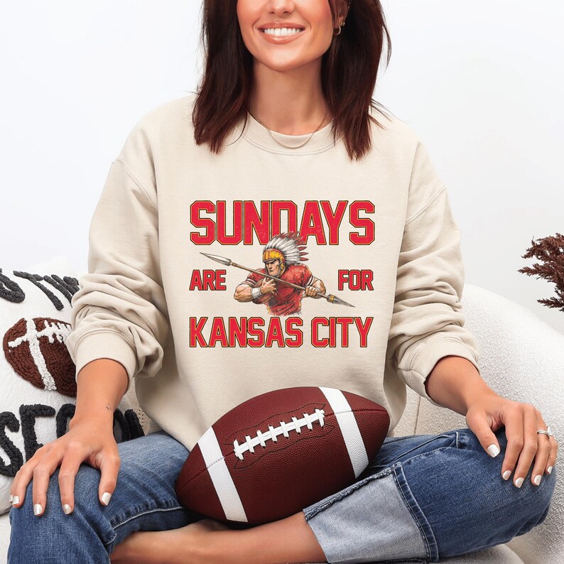A woman sitting on a couch wearing a sweatshirt with a graphic of a Native American warrior and the text "SUNDAYS ARE FOR KANSAS CITY".