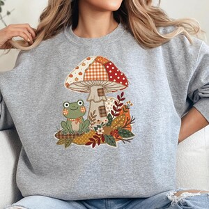 May include: A heather gray crewneck sweatshirt featuring a central graphic of a patchwork-style mushroom and a small green frog. The mushroom cap displays various patterns including pumpkins, gingham, and polka dots in orange, red, and white tones.