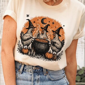 May include: A cream-colored t-shirt featuring a graphic print of three chickens wearing witch hats around a black cauldron. The design includes an orange circular background, stars, a crescent moon, pumpkins, and candles in a detailed ink-style illustration.