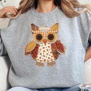 May include: A heather gray crewneck sweatshirt featuring a large, central applique of an owl. The owl is constructed from various fabric patches in shades of orange, brown, and cream, including patterns of pumpkins, plaid, and polka dots.