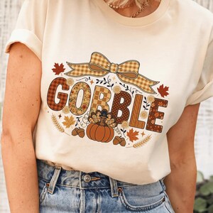 May include: A cream-colored t-shirt featuring a graphic print with the word GOBBLE in large, patterned letters. Above the text is a tan plaid bow. Below, a turkey wearing a hat sits on a pumpkin, surrounded by autumn leaves, acorns, and wheat.