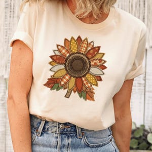 May include: A cream-colored short-sleeve t-shirt featuring a large sunflower graphic on the front. The sunflower petals are composed of various fabric-like patterns in shades of orange, yellow, brown, and green, with a dark brown center.