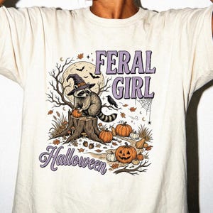 May include: A cream-colored t-shirt featuring a graphic of a raccoon wearing a witch hat perched on a tree stump. The design includes pumpkins, a full moon, bats, and the text FERAL GIRL Halloween in a purple font with a distressed texture.