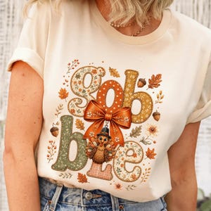 May include: A cream-colored short-sleeve t-shirt featuring a graphic print with the text "Gobble" in large, patterned letters. The design includes an orange bow, a turkey wearing a pilgrim hat, and various autumn leaves and acorns.