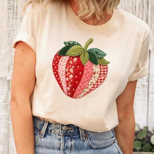 May include: A cream-colored t-shirt featuring a large, central strawberry graphic. The strawberry is composed of various red and pink fabric patches with floral, gingham, and polka dot patterns, topped with green felt leaves and a stem.