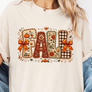 May include: A cream-colored t-shirt featuring a graphic print of the word FALL. Each letter is styled with different patterns including pumpkins, floral motifs, and a brown gingham check. Two orange bows are attached to the F and L letters.