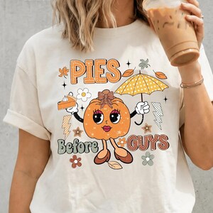 May include: A cream-colored t-shirt featuring a graphic of an orange pumpkin character with a bow, holding a slice of pie and a yellow umbrella. The text reads "PIES Before GUYS" in a retro font, surrounded by small autumn leaves, flowers, and stars.