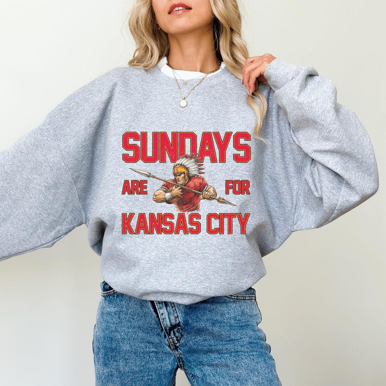 A woman wearing a grey sweatshirt with a graphic design that says "SUNDAYS ARE FOR KANSAS CITY" and features an illustration of a Native American wearing a red shirt.