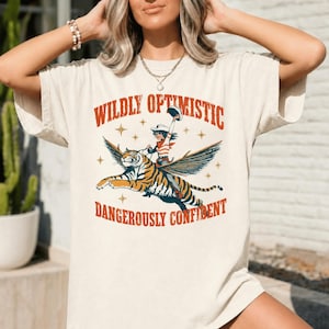 May include: A cream-colored short-sleeve t-shirt featuring a graphic print of a person in a cowboy hat riding a winged tiger. The text "WILDLY OPTIMISTIC" appears above the illustration, and "DANGEROUSLY CONFIDENT" is printed below in orange letters.