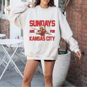 A woman wearing a white sweatshirt with a graphic design that says "SUNDAYS ARE FOR KANSAS CITY" and features an illustration of a person in a red outfit.