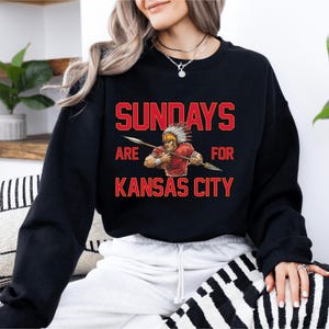 A woman wearing a black sweatshirt with a graphic design on it, which says "SUNDAYS ARE FOR KANSAS CITY".