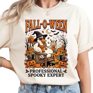 May include: A cream-colored t-shirt featuring a graphic of a white duck wearing a witch hat and heart-shaped sunglasses, sitting in a chair with a drink. The text reads FALL-O-WEEN and PROFESSIONAL SPOOKY EXPERT, surrounded by pumpkins, cats, and ghosts.