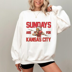 A woman wearing a white sweatshirt with a graphic design that says "SUNDAYS ARE FOR KANSAS CITY" and features an illustration of a Native American warrior.