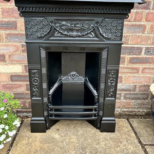 May include: A matte black cast iron fireplace insert featuring an ornate mantel shelf, a central frieze with a fruit and leaf garland, and side panels decorated with floral motifs. The interior includes a metal grate and a decorative arch.