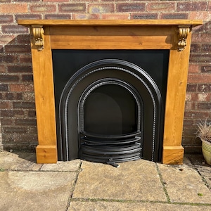 Puede incluir: Una chimenea de madera con un inserto de hierro fundido negro. La repisa está hecha de madera de color claro con tallas decorativas en las esquinas. El inserto tiene un diseño arqueado con una rejilla en la parte inferior. La chimenea está contra una pared de ladrillo.