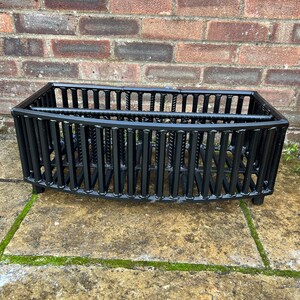 May include: A rectangular black metal fireplace grate constructed from vertical steel bars with a curved front design. The structure features a sturdy frame with a horizontal support bar across the top, designed to hold firewood securely within a hearth.