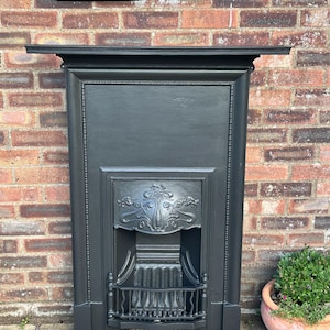 May include: A matte black fireplace surround featuring a rectangular mantelpiece, a beaded frame, and a central cast iron insert. The insert displays an intricate embossed floral motif above a metal grate with vertical bars and a curved base.
