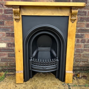 May include: A classic fireplace surround with a black cast iron firebox and a light wood frame. The mantelpiece has ornate corbels and a plain top. The firebox features an arched design and a decorative grate.