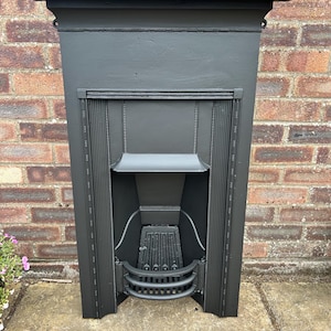 May include: A matte black cast iron fireplace insert featuring a rectangular mantel top, vertical fluted side columns with beaded detailing, a central hood, and a curved grate at the base. The structure has a solid, uniform dark finish.