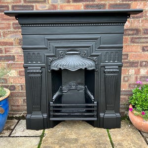 May include: A matte black cast iron fireplace surround featuring an ornate arched hood, fluted pilasters, and a central grate. The structure displays intricate relief detailing, including geometric patterns and a scalloped canopy over the hearth.