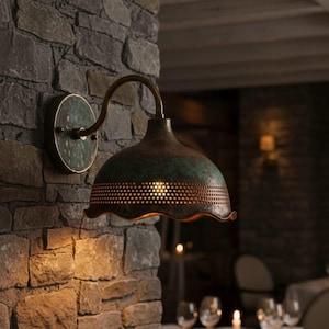 Puede incluir: Una lámpara de pared de estilo antiguo con acabado cobrizo y pátina verde. La lámpara presenta un brazo curvo y una pantalla en forma de campana con borde festoneado y pequeñas perforaciones. Está montada en una pared de piedra.