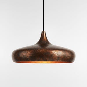 May include: A copper-coloured pendant light fitting with a hammered texture. The light has a rounded, bell-like shape with a narrow neck and is suspended from a black cord. The interior of the light fitting has a warm, golden glow.