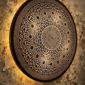 Peut inclure: Applique murale ronde en métal noir avec un motif floral complexe. La lumière est allumée, créant une lueur chaude sur un mur texturé et vieilli. Le design rappelle le décor marocain ou du Moyen-Orient.