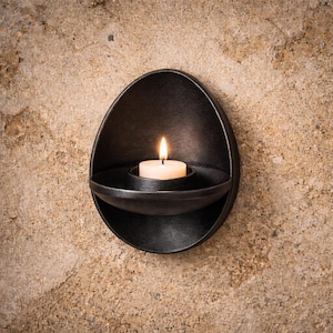 Könnte beinhalten: Ein schwarzer, an der Wand befestigter Kerzenhalter in Tropfenform. Eine brennende weiße Kerze befindet sich in der Mitte und wirft einen warmen Schein. Der Halter ist aus Metall und an einer strukturierten, beigefarbenen Wand befestigt.