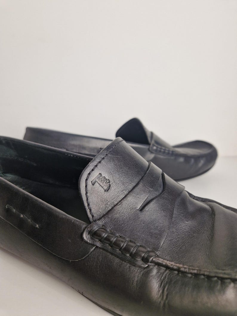 May include: Close-up of two black leather loafers. The loafers have a classic design with a penny strap and a stitched vamp. The leather appears smooth and the shoes are in good condition. The background is a plain white.