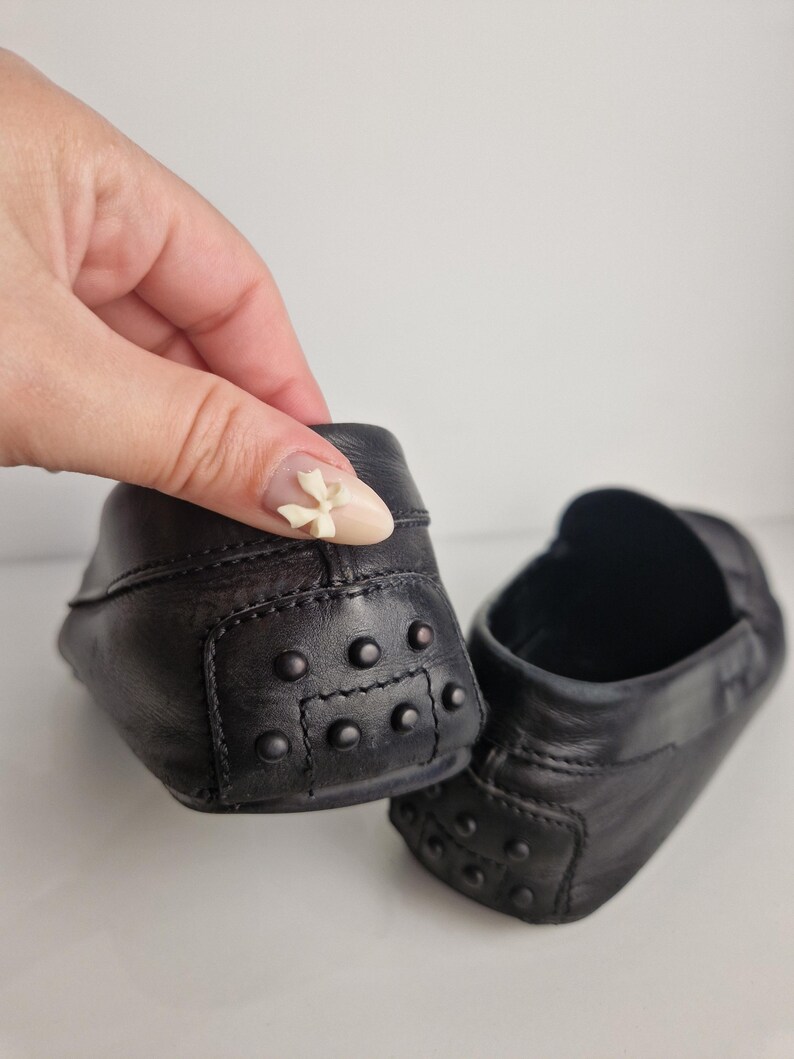 May include: A pair of black leather baby loafers. The shoes have a classic design with a rounded toe and rubber pebble detailing on the heel. The shoes are made of a soft, supple leather.
