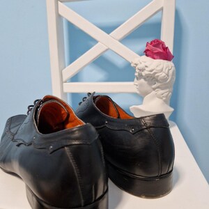 Puede incluir: Un par de zapatos de vestir de cuero negro con costuras decorativas y puntera cuadrada. Los zapatos se exhiben sobre una superficie blanca con una silla blanca y un busto con una flor rosa.