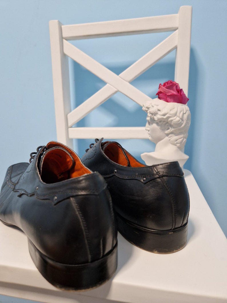 Puede incluir: Un par de zapatos de vestir de cuero negro con costuras decorativas y puntera cuadrada. Los zapatos se exhiben sobre una superficie blanca con una silla blanca y un busto con una flor rosa.