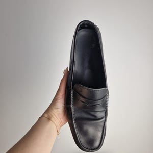 May include: Black leather penny loafers. The shoes have a classic design with a rounded toe and a strap across the vamp. The loafers are held up against a white background.