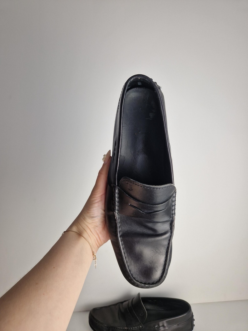 May include: Black leather penny loafers. The shoes have a classic design with a rounded toe and a strap across the vamp. The loafers are held up against a white background.