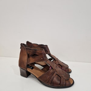 May include: A pair of brown leather sandals with an open toe design. The sandals have a low heel and a zipper closure at the back. The brand name "KKA" is visible on the side. The sandals are displayed on a white surface.