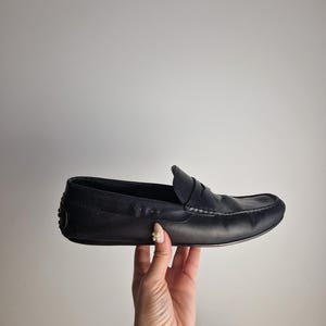 May include: A black leather loafer with a rounded toe and a penny keeper strap. The shoe is held up by a hand against a white background. The shoe has a stitched design along the top.