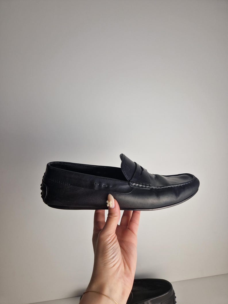 May include: A black leather loafer with a rounded toe and a penny keeper strap. The shoe is held up by a hand against a white background. The shoe has a stitched design along the top.