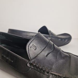May include: Close-up of two black leather loafers. The loafers have a classic design with a penny strap and a stitched vamp. The leather appears smooth and the shoes are in good condition. The background is a plain white.