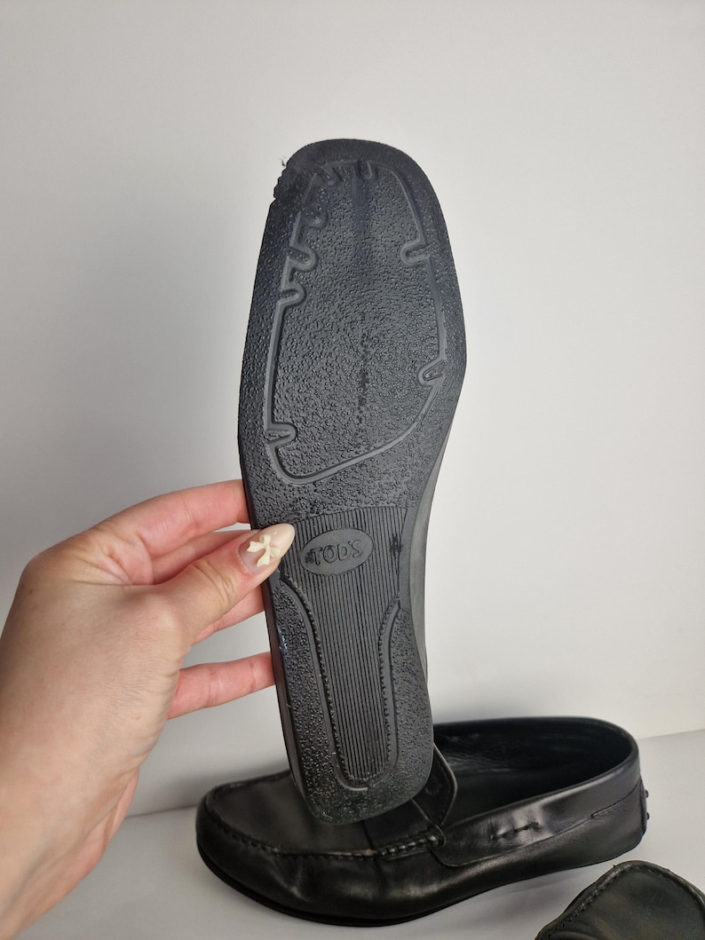 May include: Black leather loafers with a rubber sole. The sole is textured with a pattern of grooves and the brand name "TOD'S" is visible. The shoe is held up to show the sole.