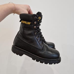 Puede incluir: Botas de trabajo de cuero negro con el nombre de la marca "Caterpillar" en letras amarillas. Las botas tienen un dise&ntilde;o con cordones, un cuello acolchado y una suela gruesa y resistente. Las botas est&aacute;n sujetas por una mano.
